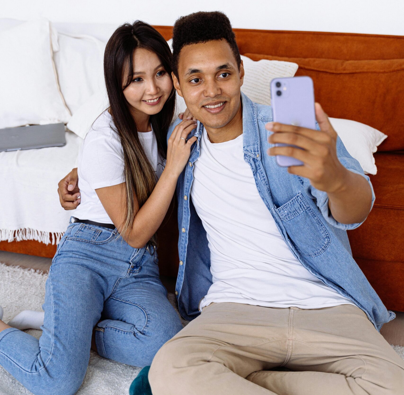 Interracial couple sitting indoors capturing a selfie on a smartphone.