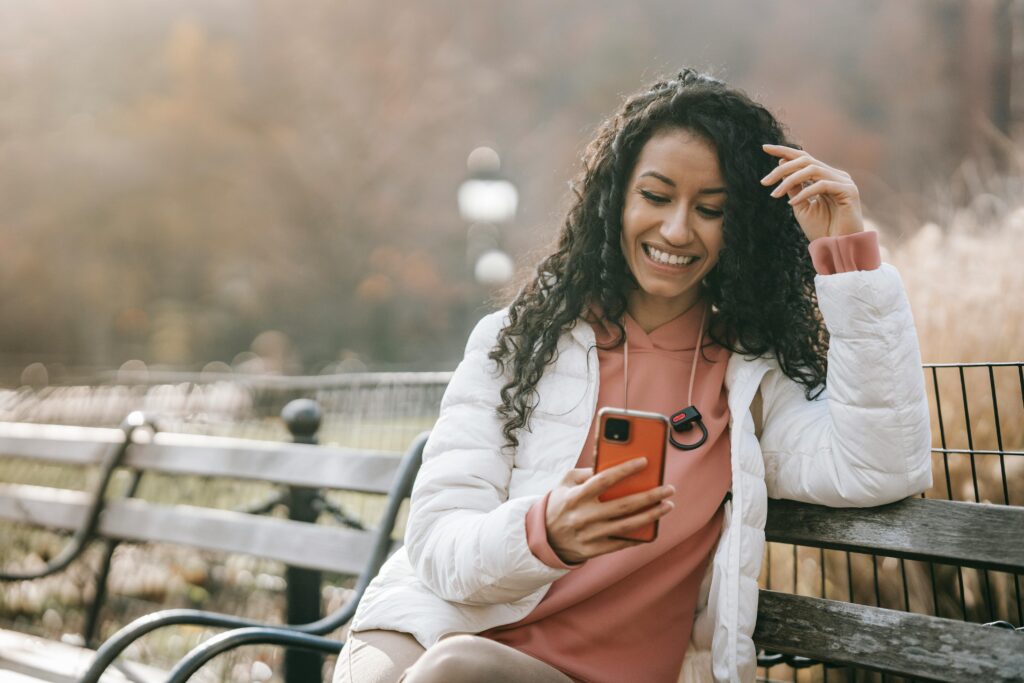 A cheerful woman enjoying a sunny autumn day, sitting on a bench and browsing her smartphone in the park.